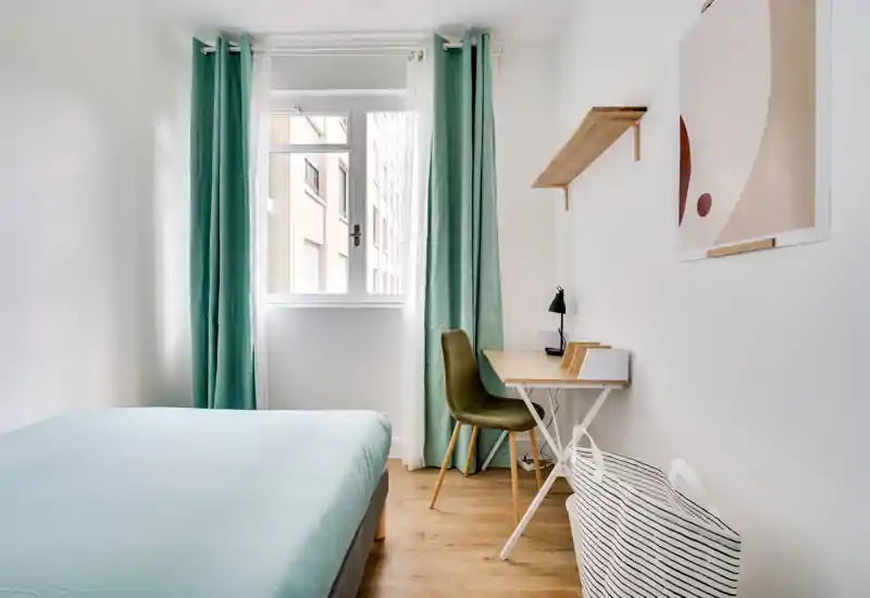 Bright, minimal bedroom with a neatly made bed, pale blue linens and a small wooden desk by the window—simple, modern styling and good natural light.