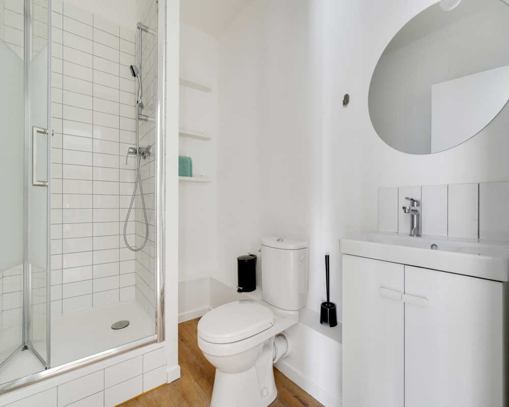 Bright, modern bathroom with a tiled shower enclosure, toilet, sink cabinet and round mirror — clean white finishes and wood-effect flooring.