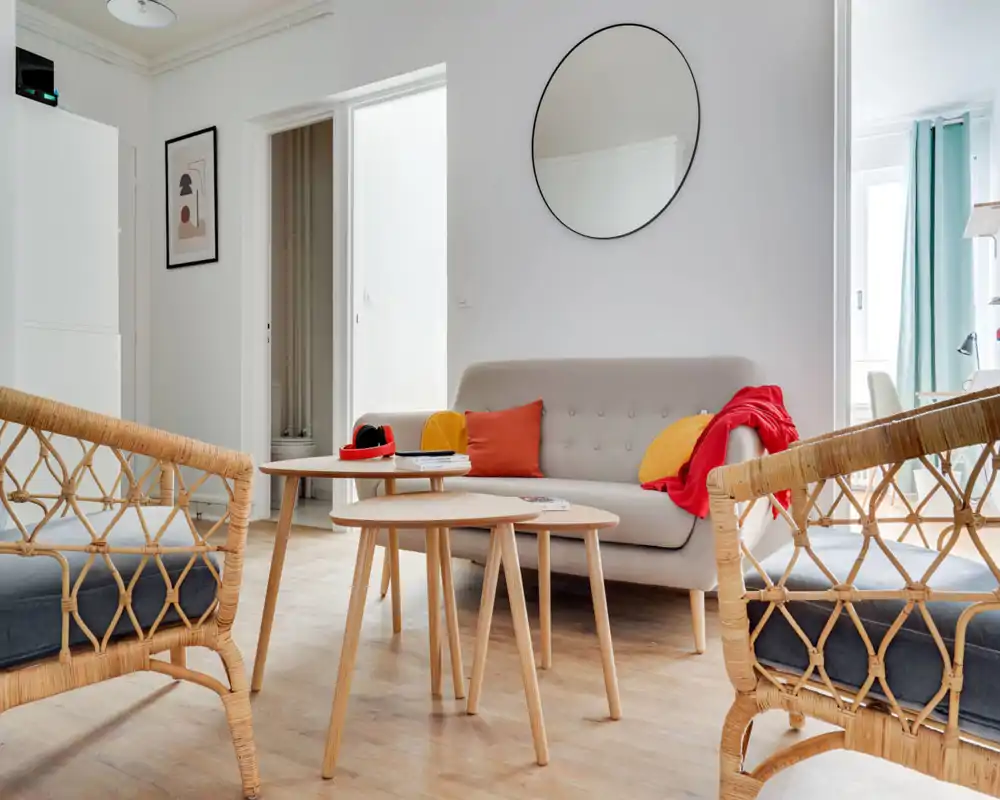 Bright, stylish living room with a small sofa, colorful cushions and natural-wood nesting coffee tables; rattan armchairs add warmth and a large round mirror enhances the sense of space.