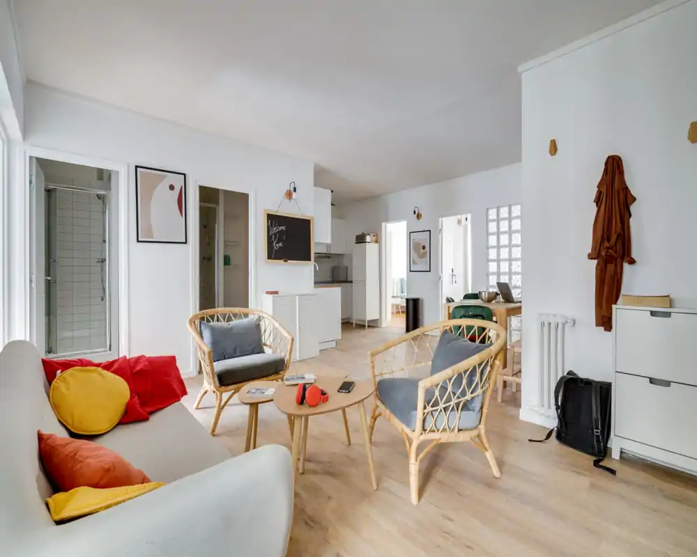 Bright, modern living area with a cozy seating arrangement including a sofa, rattan armchairs and a small coffee table; light wood floors and clean white walls create an airy feel.