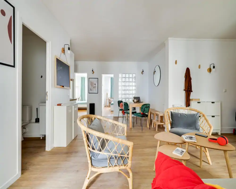 Bright, stylish open living area with rattan seating, a small coffee table and a dining nook in the background. Natural light and warm wood floors create a welcoming, modern atmosphere.
