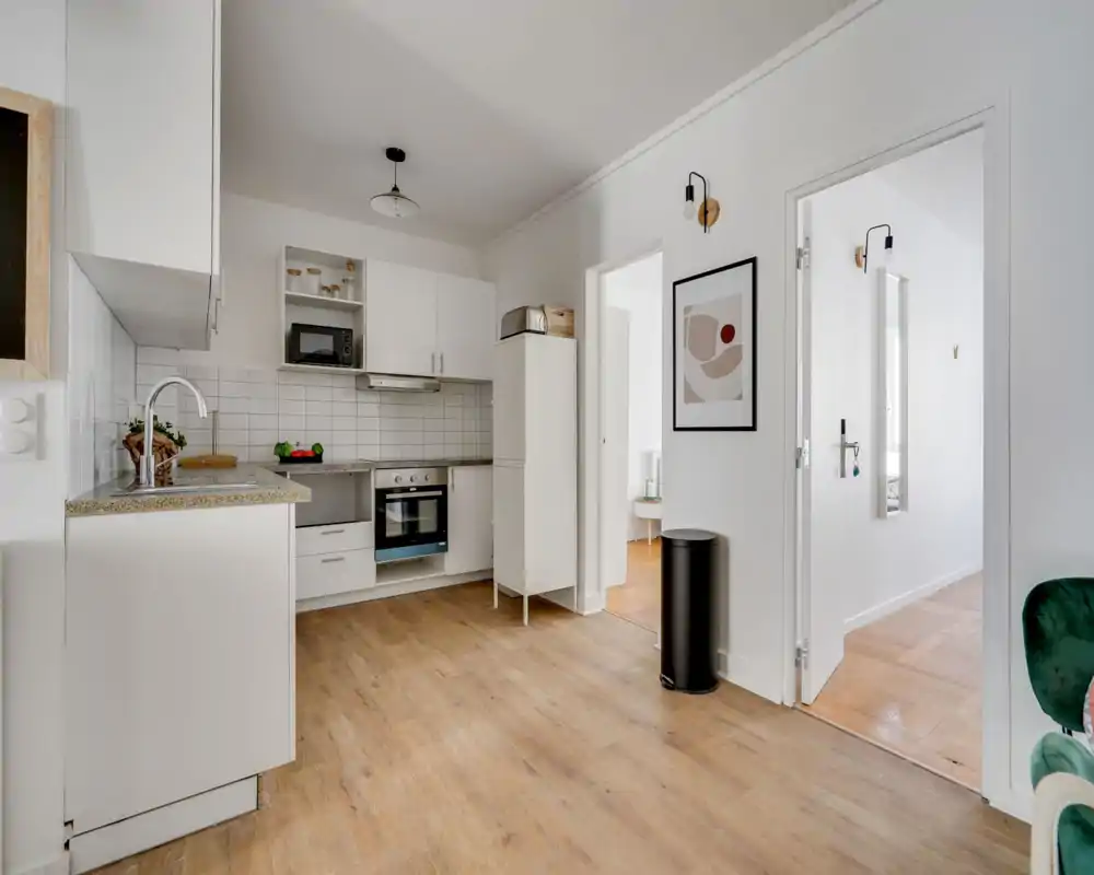 Bright modern kitchen with white cabinetry, tiled backsplash, integrated oven and countertop sink. Clean, minimal styling with wood-effect flooring and small decorative accents.