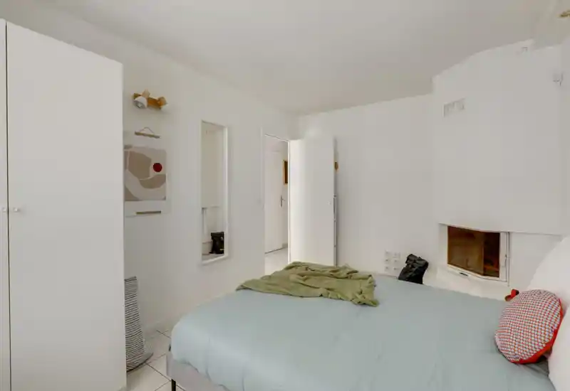 Bright, minimal bedroom with a centrally visible double bed dressed in pale linens, a small fireplace at the corner and simple white storage; calm modern styling ideal for a rental listing.