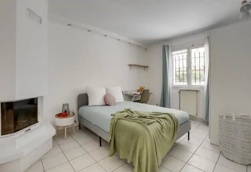 Bright, minimalist bedroom with a double bed dressed in pale linens and a soft green throw, a small bedside table, and a desk by the window providing natural light — ideal for a calm, modern rental.