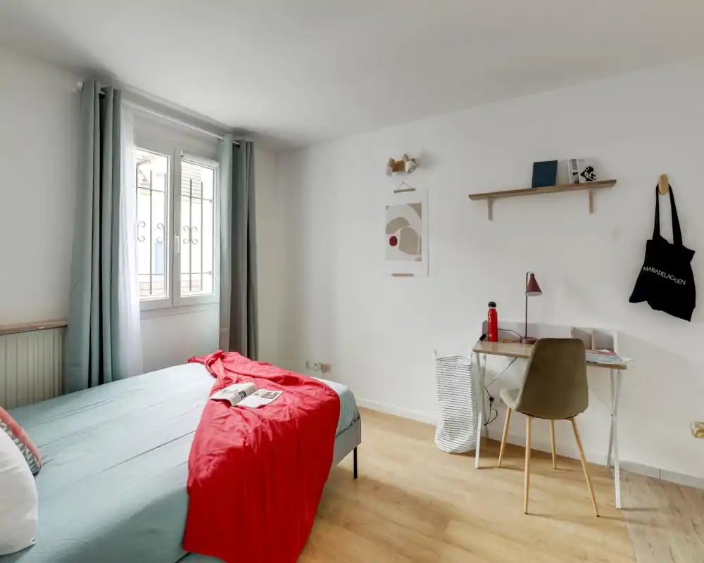 Bright, minimalist bedroom with a full bed dressed in soft linens and a red throw, a window with curtains letting in natural light, and a small desk area—ideal for a comfortable stay or working corner.