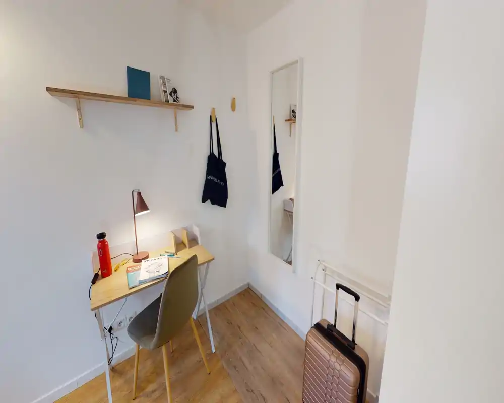 Small bedroom corner with a compact desk, chair, lamp and luggage by the wall; bright white walls and wooden flooring create a simple, functional space.