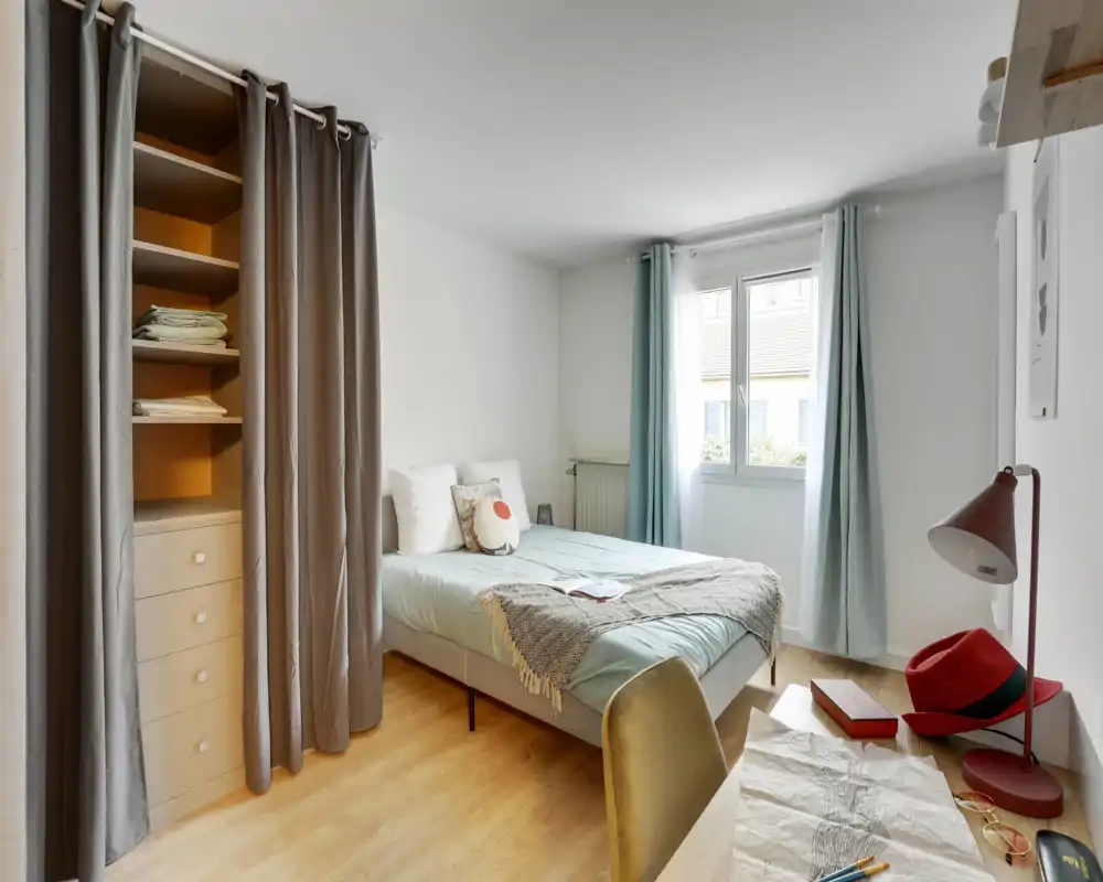 Chambre lumineuse et bien décorée avec un lit double, linge doux, coussin décoratif et un bureau; la lumière de la fenêtre et les rideaux pastel apportent une ambiance chaleureuse.