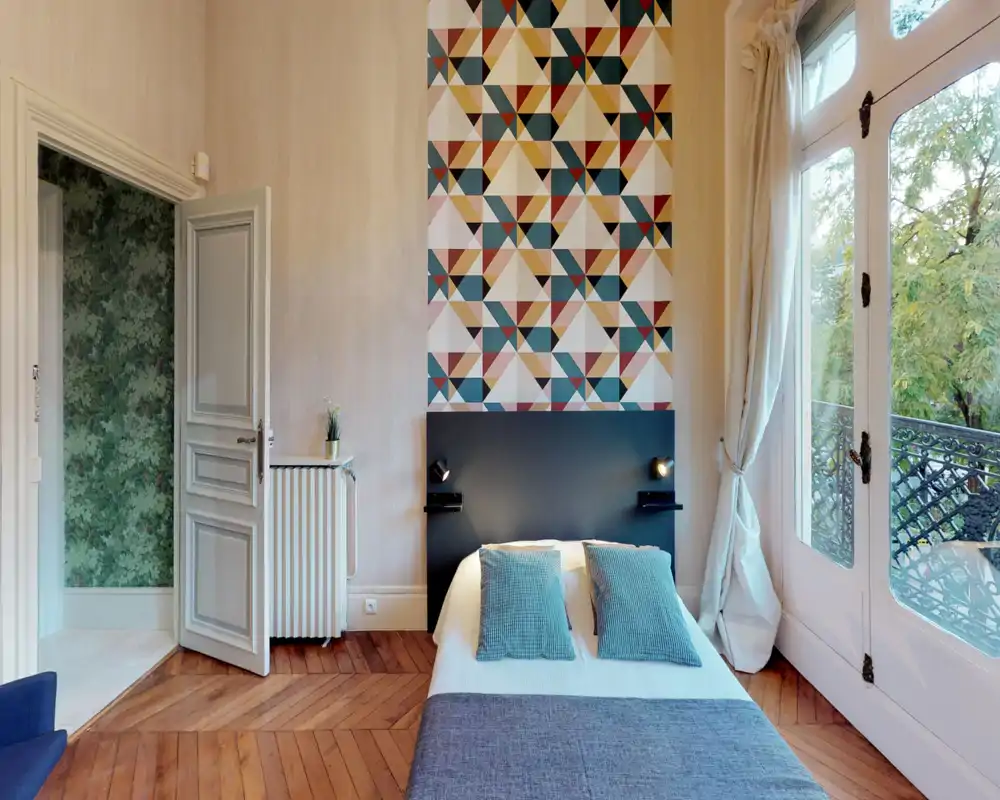 Stylish, bright bedroom with a patterned accent wall, a centered double bed with two teal cushions, and large French windows opening to greenery — ideal for rental photos highlighting light and design.