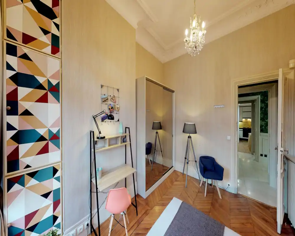 Bright, stylish bedroom with wooden herringbone floor, patterned built-in wardrobe and a visible bed corner. The room includes a small workspace, tripod floor lamps and a mirrored wardrobe that enhances the sense of space.