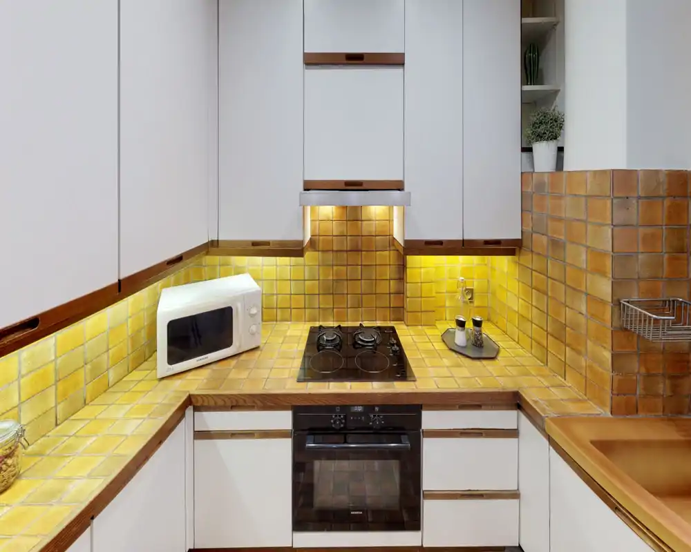 Compact, well-equipped kitchen with warm yellow tile backsplash, under-cabinet lighting, built-in oven and four-burner cooktop — ideal for cozy meal prep.