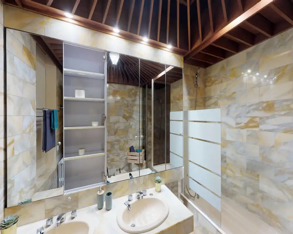 Well-lit marble bathroom with a double sink counter, large mirrored cabinet, bathtub with glass divider and overhead shower — modern, warm finishes and wooden ceiling beams add character.