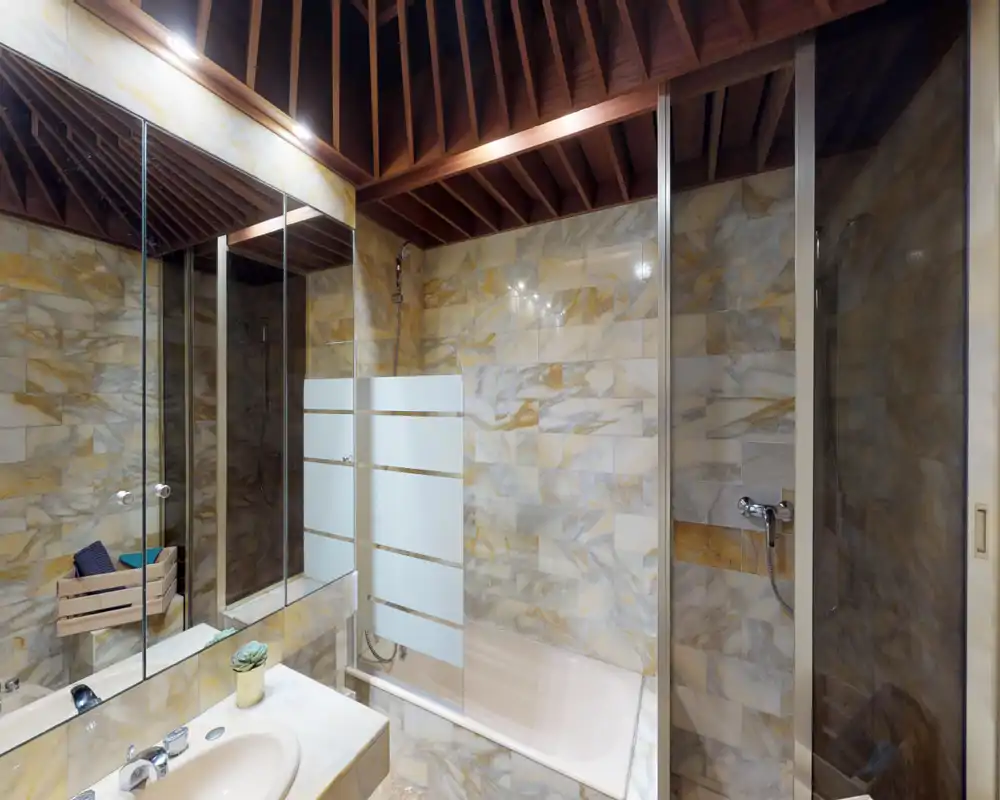 Bright, stylish bathroom with marble-effect tiles, a bathtub with glass partition and a wooden slatted ceiling; vanity with sink and mirrored cabinet.
