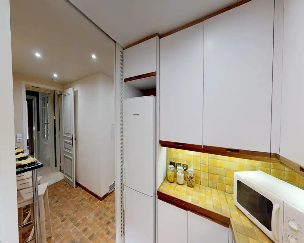 Compact, well-lit kitchen corner with warm yellow tiled countertop, white cabinetry, refrigerator and microwave — practical and cozy for a small apartment.