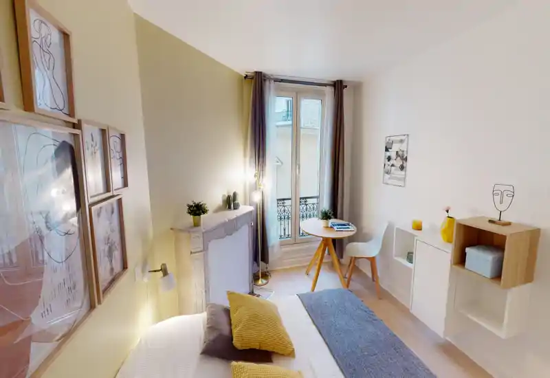 Chambre lumineuse et soignée avec un lit double bien fait, coussins décoratifs et plaid. Lumière naturelle par de grandes fenêtres, petite table ronde près de la fenêtre et décor mural élégant pour une ambiance moderne et cosy.