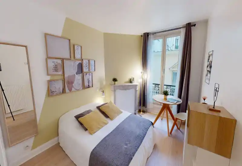 Chambre lumineuse et stylée avec un lit double habillé de linge neutre, coussins décoratifs et jeté texturé. Décorations murales encadrées, petite table près de la fenêtre, lampe sur pied et belle lumière naturelle d'une fenêtre balcon.