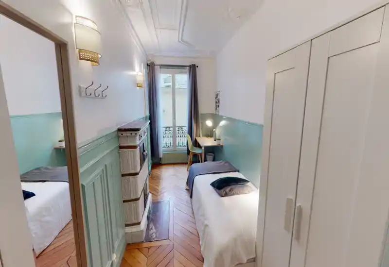Chambre étroite et lumineuse avec parquet, moulures décoratives et un lit simple avec coussins ; un petit bureau près de la fenêtre offre un coin travail chaleureux.