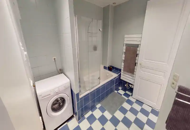 Salle de bain propre et lumineuse avec baignoire et pare-douche en verre, carrelage au sol et aux murs, machine à laver et radiateur porte-serviettes.