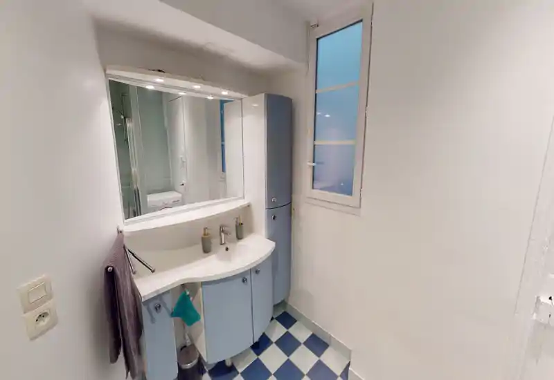 Salle de bain compacte et moderne avec lavabo courbé, miroir éclairé et sol à carreaux bleu et blanc — lumineuse et bien rangée.