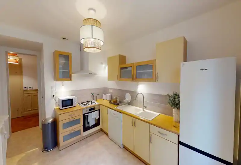 Cuisine compacte et lumineuse avec plans de travail en bois, four encastré, plaque à gaz, lave-vaisselle et grand réfrigérateur. Éclairage chaleureux et agencement soigné, parfait pour une annonce locative.