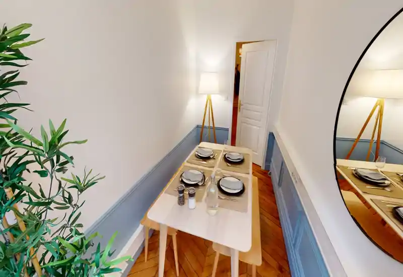 Espace repas étroit dressé pour quatre personnes avec table en bois, sets de table et lampe chaleureuse; décoration contemporaine avec miroir rond et plante d’intérieur.