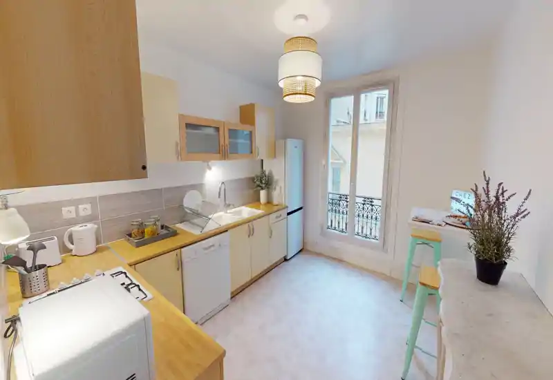 Cuisine moderne et lumineuse avec plans de travail en bois, placards hauts, grand réfrigérateur et lave-vaisselle. La grande fenêtre et l'éclairage suspendu apportent une ambiance chaleureuse.