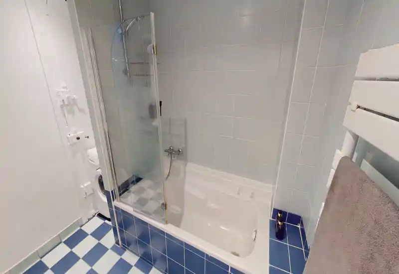 Salle de bains propre et bien éclairée avec baignoire-douche, paroi de douche en verre dépoli, habillage bleu autour de la baignoire et sol à carreaux.