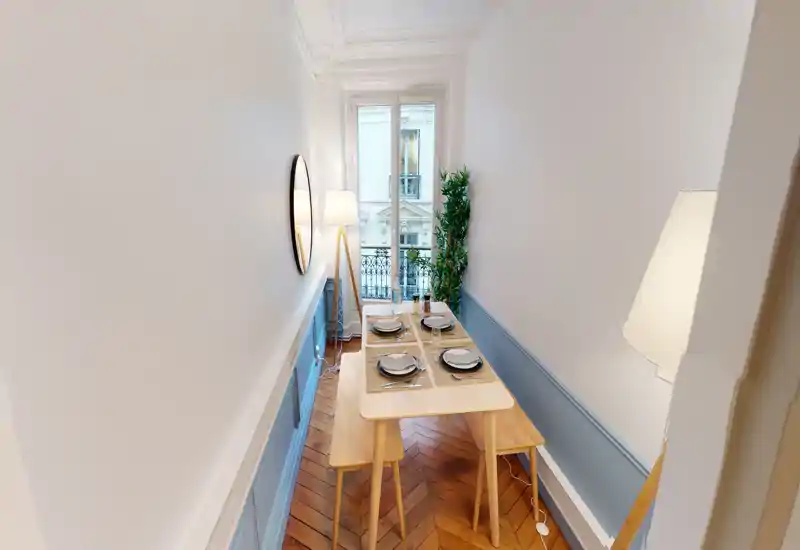 Coin repas étroit avec table en bois dressée pour quatre, banquettes et lumière douce. Grande fenêtre et murs clairs offrent une ambiance aérée et élégante, parfaite pour des repas intimes.