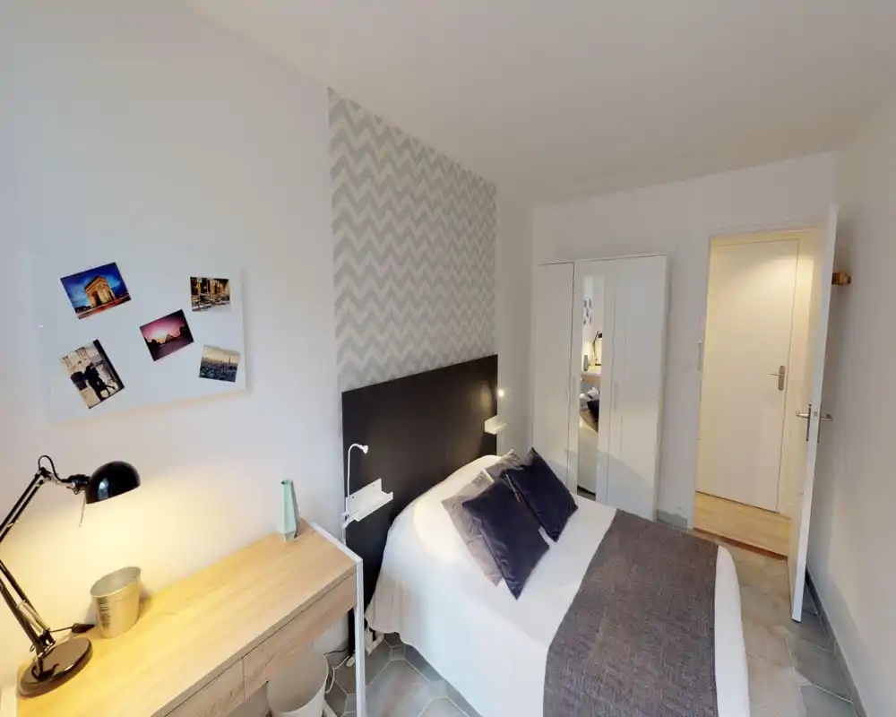 Comfortable, well-lit bedroom with a double bed, decorative pillows and a chevron-accent wall. A desk with lamp and a mirrored wardrobe complete the functional layout, ideal for short-term rentals.