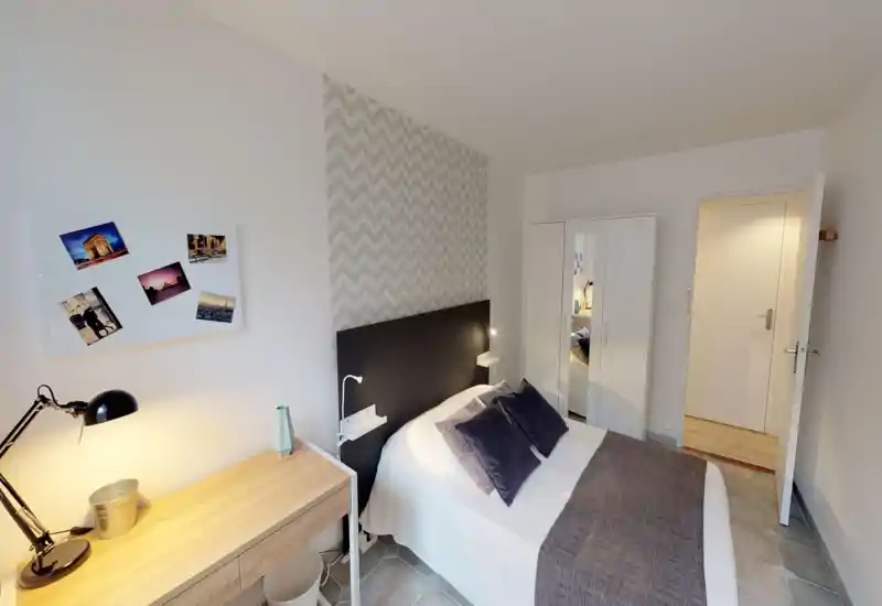 Comfortable, well-lit bedroom with a double bed, decorative pillows and a chevron-accent wall. A desk with lamp and a mirrored wardrobe complete the functional layout, ideal for short-term rentals.