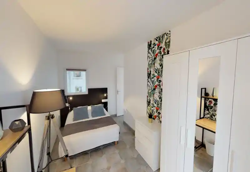 Chambre moderne et lumineuse avec un lit double, lampes de chevet, une bande de papier peint décoratif et une armoire blanche avec porte miroir. Décor soigné avec coussins, lampadaire et un petit bureau visible dans le miroir.