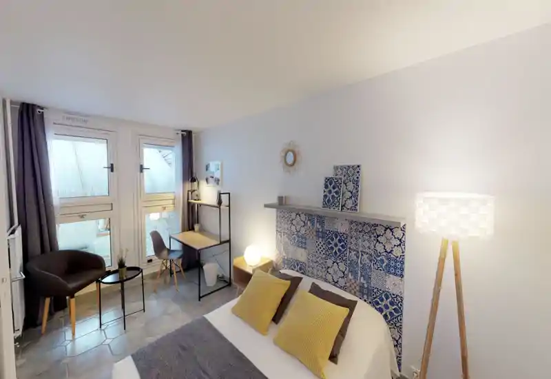 Chambre lumineuse et soignée avec une tête de lit à motifs, deux coussins jaunes et un éclairage en couches. Un bureau compact et un coin salon près de grandes fenêtres rendent la pièce fonctionnelle pour le repos et le travail.