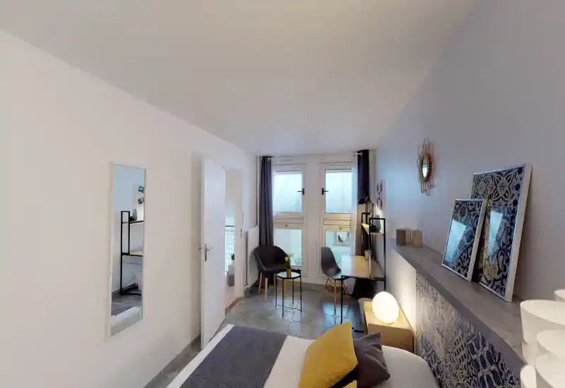 Chambre claire et bien aménagée avec un lit visible au premier plan, coussins confortables et lampes de chevet ; un petit bureau et des sièges près des fenêtres créent un coin travail pratique.