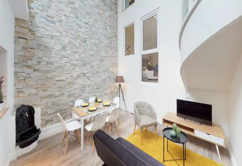 Espace de vie lumineux et moderne avec mur en pierre, table à manger pour six, assises confortables et meuble TV — mise en scène soignée pour location.