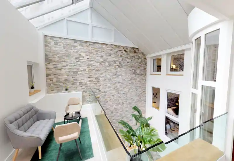 Espace mezzanine lumineux avec un canapé gris moderne, deux fauteuils beiges et une petite table basse sur un tapis vert ; la verrière et le mur en pierre apportent une ambiance élégante et accueillante.