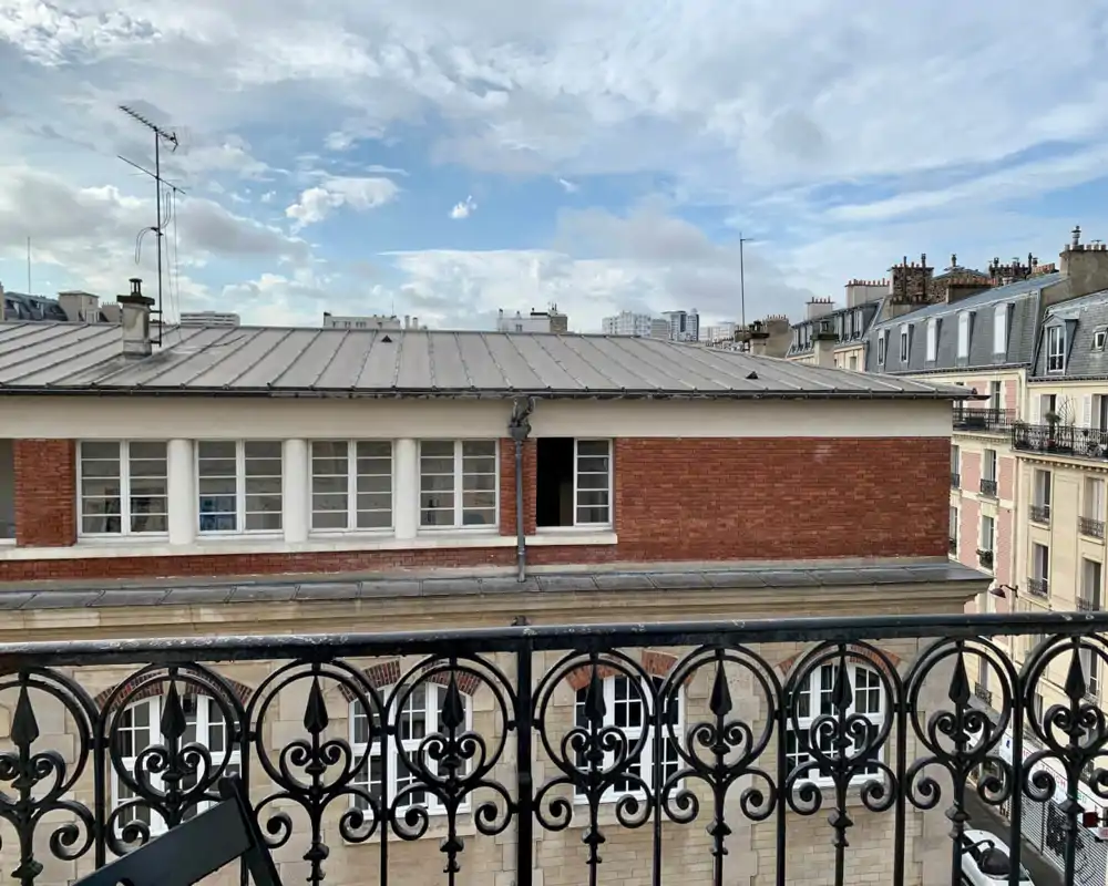 Petit balcon avec une balustrade en fer forgé décorative et vue sur les toits voisins sous un ciel lumineux — parfait pour un café du matin.