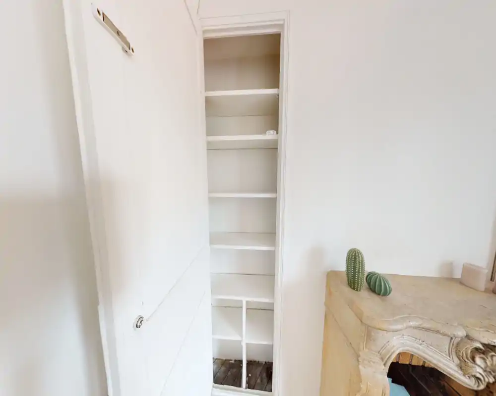 Petite zone d'entrée avec un placard étroit à étagères et une console décorative. Murs blancs et sol en bois offrent un espace d'accueil propre et lumineux.