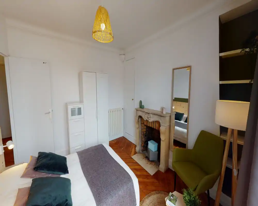 Chambre lumineuse et décorée avec un grand lit, des coussins décoratifs, une cheminée ancienne et un fauteuil cosy près de la fenêtre — idéale pour un séjour confortable.