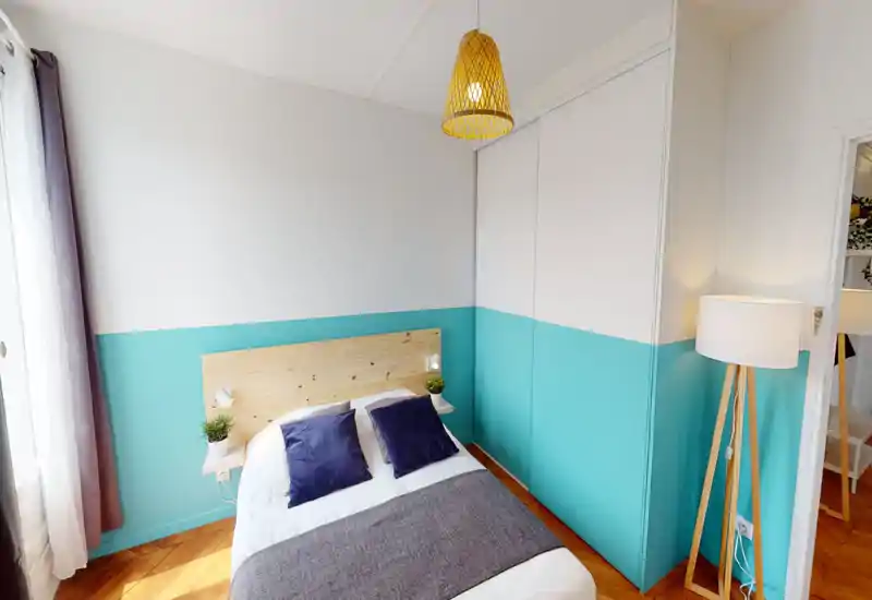 Chambre lumineuse et aménagée avec lit double, tête de lit en bois, bande murale turquoise et lumière naturelle venant de la fenêtre. Coussins assortis, petites plantes et lampadaire donnent une ambiance chaleureuse et moderne.