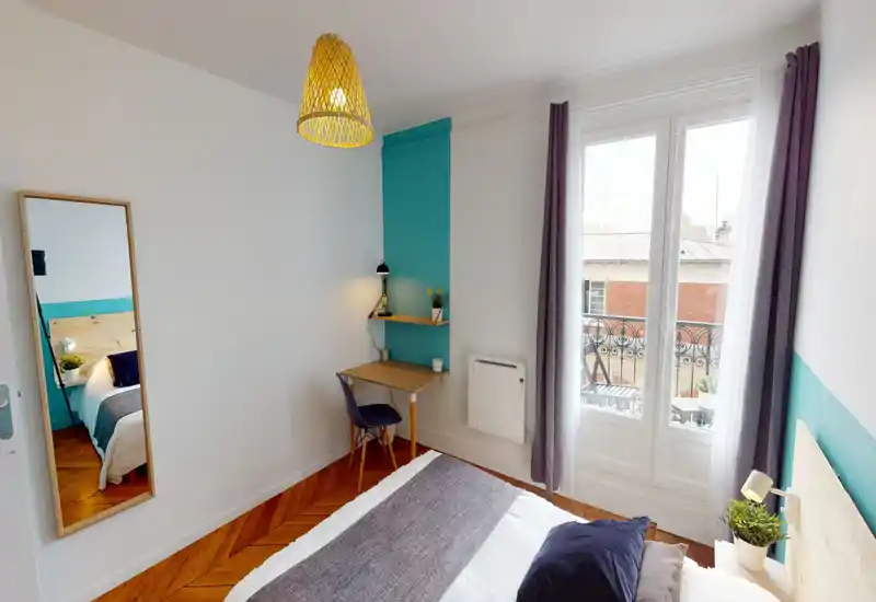 Chambre lumineuse et chaleureuse avec un lit double, tête de lit en bois clair et textiles doux. Un petit bureau près de la fenêtre et des plantes décoratives rendent l'espace fonctionnel et accueillant.