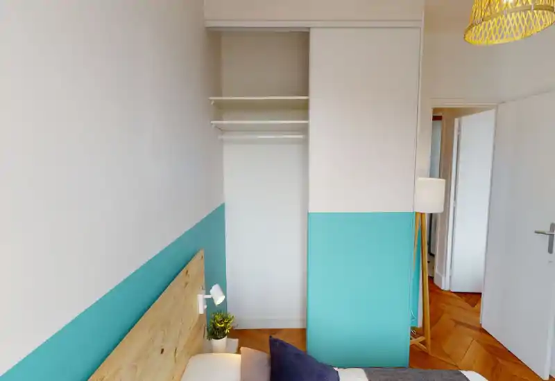 Chambre moderne et lumineuse avec tête de lit en bois, lampe de chevet et mur d'accent turquoise. La photo met en avant l'espace lit et un placard ouvert.