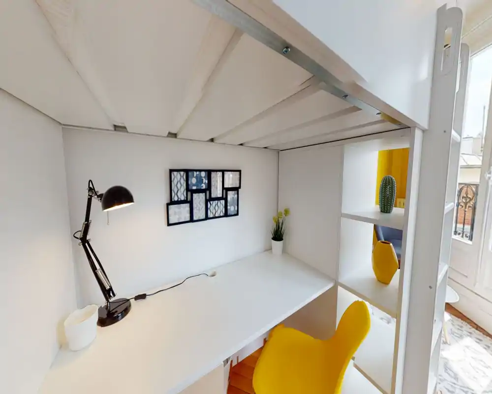 Espace nuit compact avec coin bureau sous un lit mezzanine : bureau blanc épuré, lampe de bureau noire et chaise jaune vive. La lumière naturelle et quelques décorations (plante et cadres) offrent un coin travail moderne et lumineux.