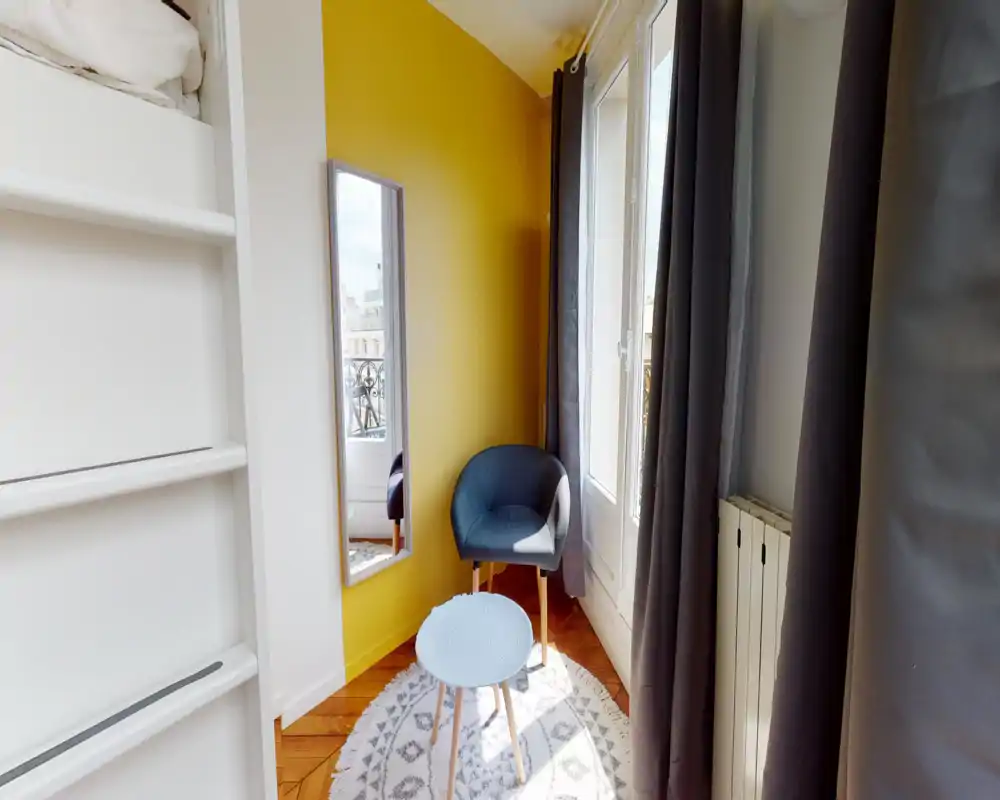 Coin salon lumineux et chaleureux avec un fauteuil, une petite table ronde et une grande glace près d'une fenêtre ensoleillée — idéal pour lire ou se détendre.