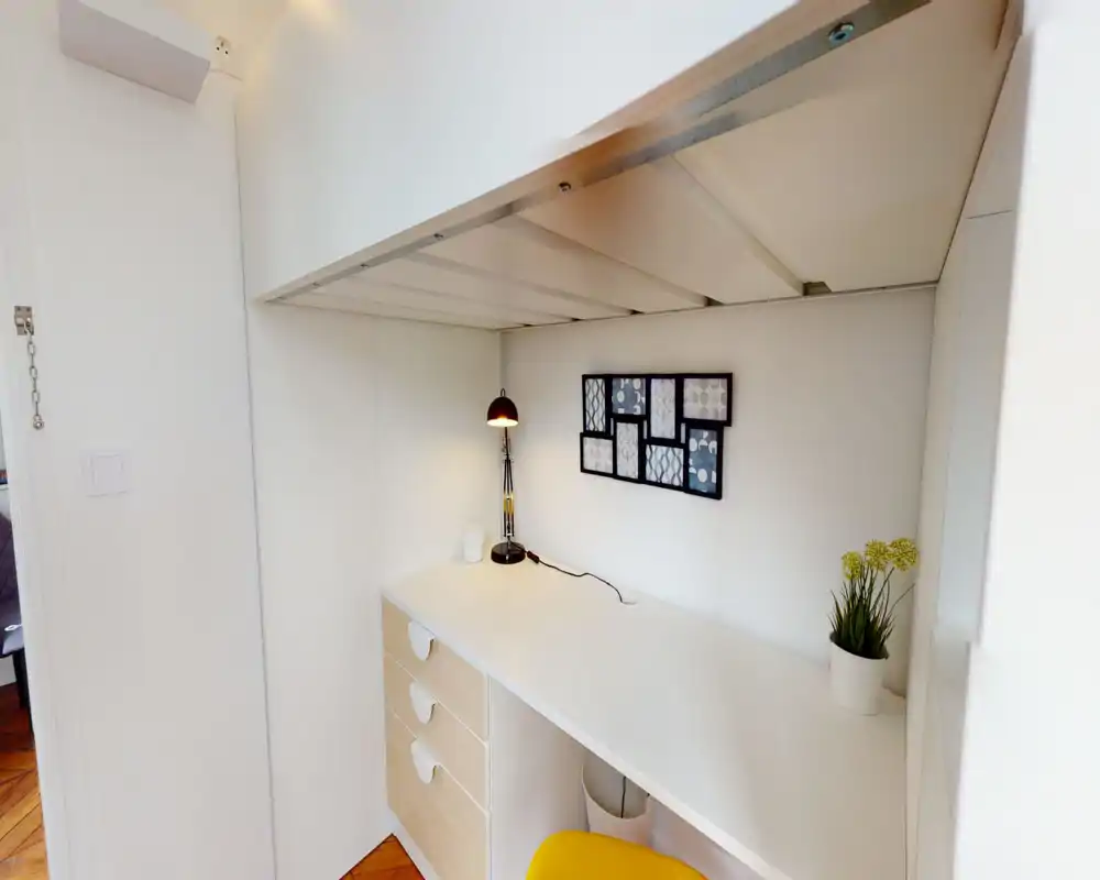 Coin de chambre compact avec lit mezzanine et bureau intégré. L'espace de travail est lumineux et soigné, avec une lampe de bureau, des cadres muraux et une petite plante en pot.