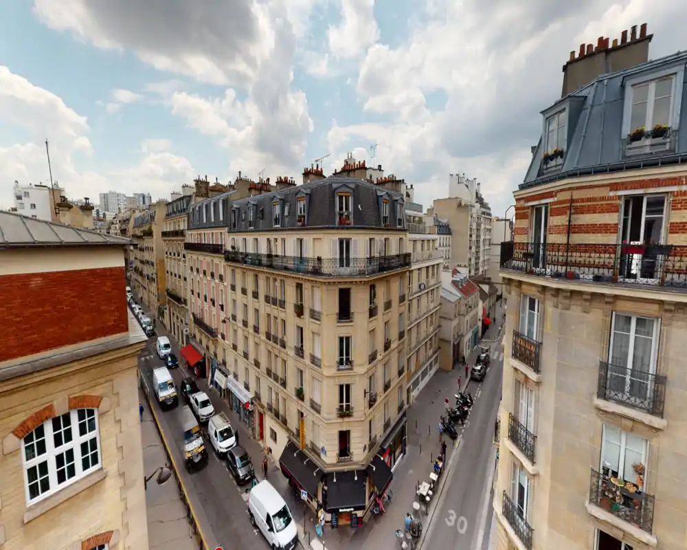 Vue extérieure urbaine au niveau de la rue montrant des façades d'immeubles à la parisienne, toits et un café d'angle — ciel lumineux et animation de rue pour une belle scène citadine.