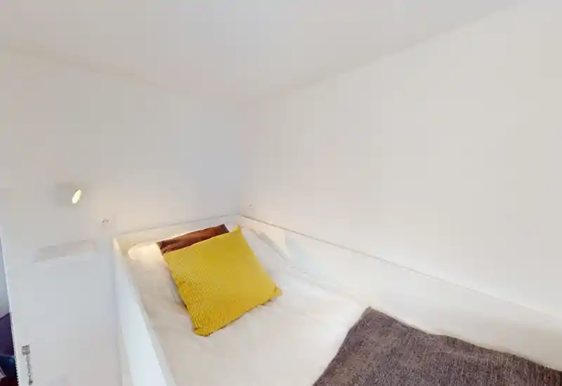 Coin chambre lumineux et minimaliste avec un lit simple, un coussin jaune et du linge blanc ; murs épurés et éclairage doux pour une atmosphère calme et aérée.