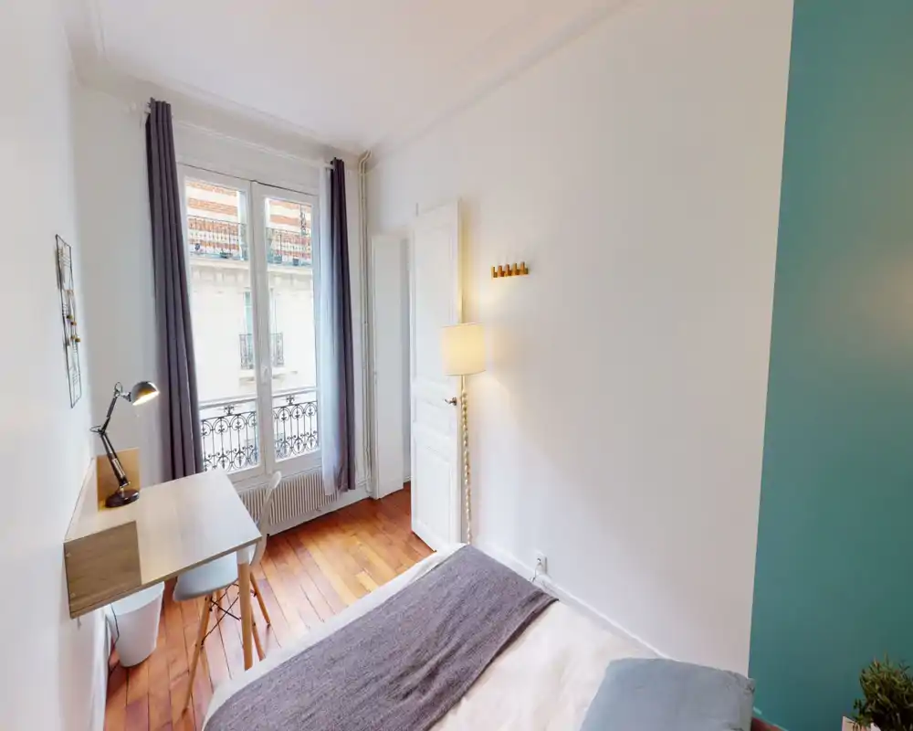 Bright, compact bedroom with a comfortable bed, wooden floors and a small work desk by the window. The large window and floor lamp provide pleasant natural and artificial light for a cozy stay.