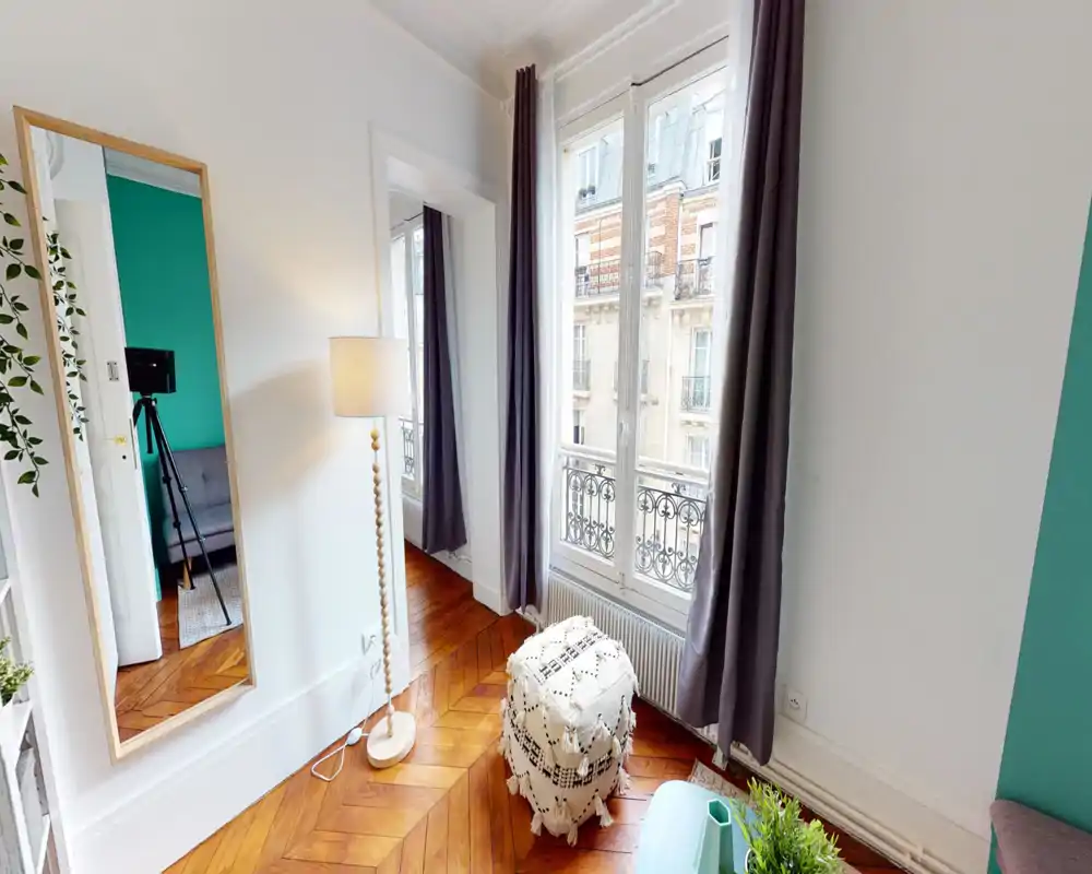 Bright, stylish living area with a large window, wooden parquet flooring, cozy pouf and floor lamp—comfortable space with modern decorative touches.