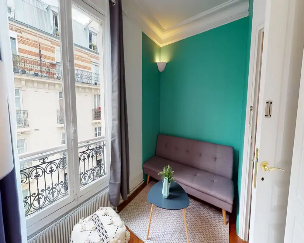 Small, bright living area with a compact sofa against a teal accent wall, a coffee table and large windows that flood the room with natural light — ideal for a cozy city apartment.