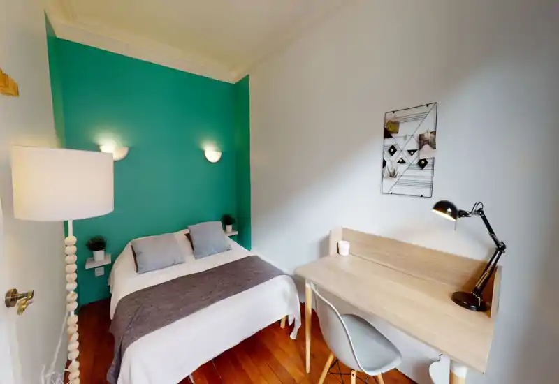 Bright, well-styled bedroom with a double bed against a teal accent wall, bedside lighting and a compact wooden desk — comfortable and practical for both rest and work.