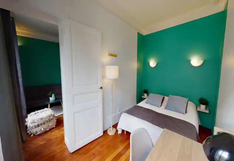 Bright, stylish bedroom with a double bed against a teal accent wall, bedside shelves with plants, warm wooden floors and soft wall lamps creating a cozy atmosphere.